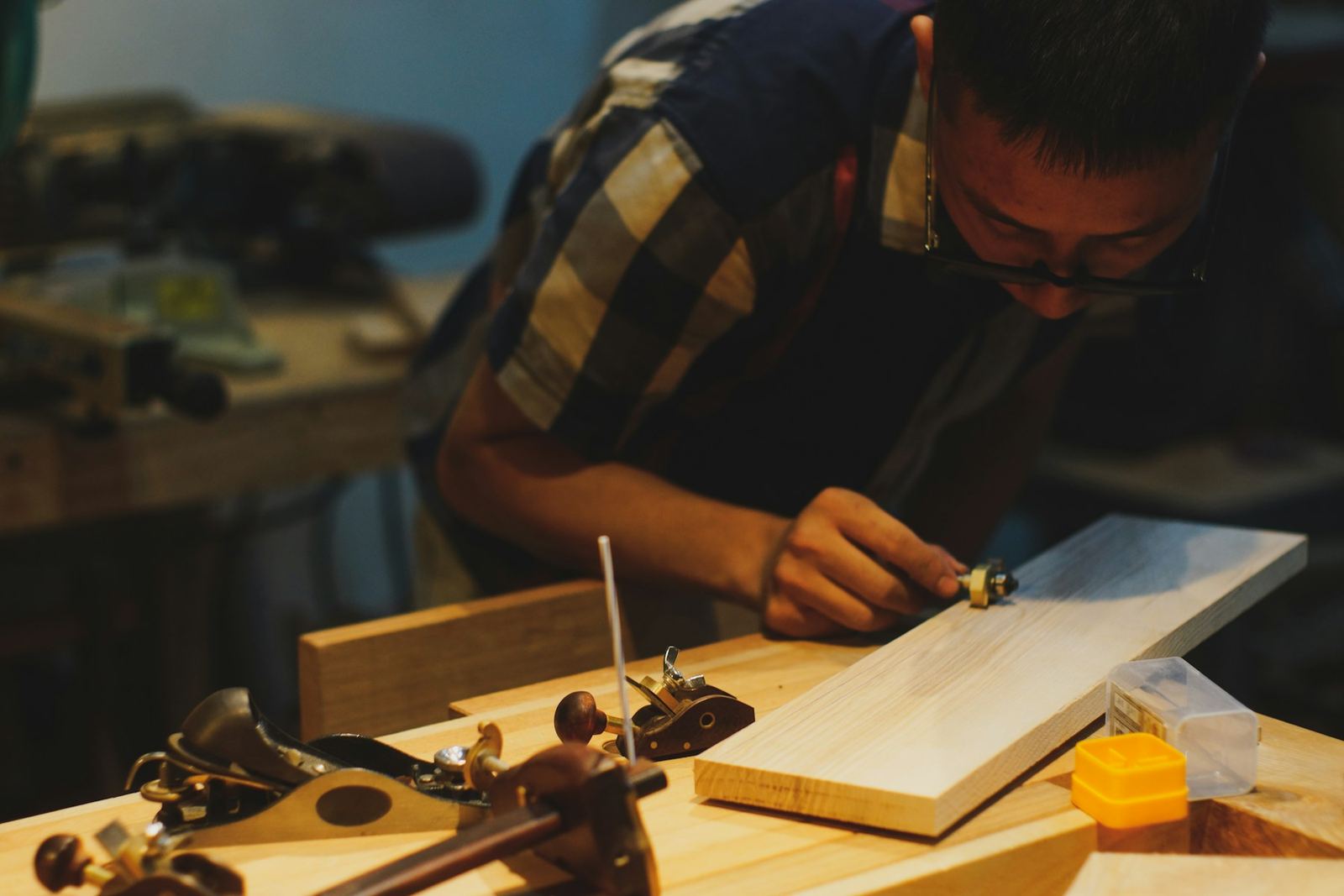 Carpenter in a plaid shirt planing a pale board at a workbench lined with hand planes